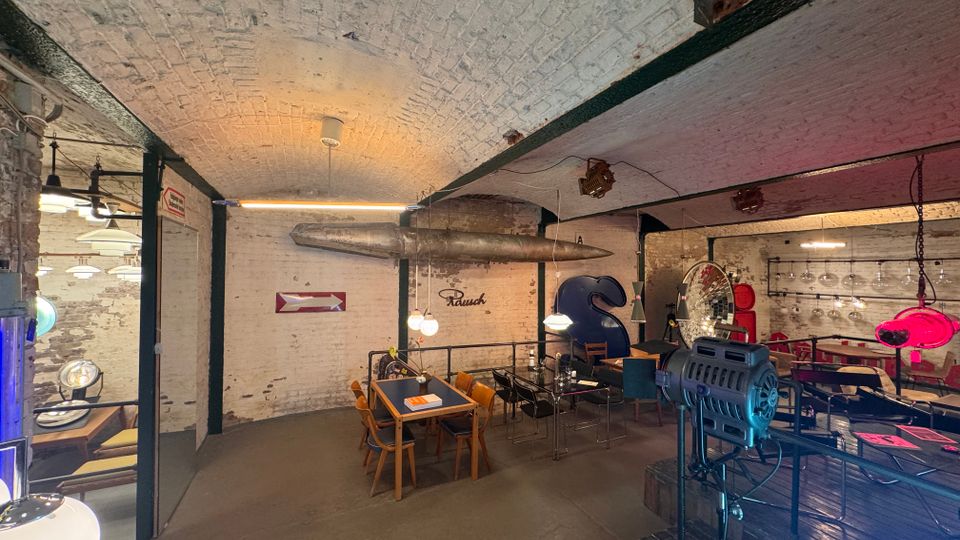 Industrie-inspiriertes Interieur in einem Raum mit gewölbter Ziegeldecke, sichtbaren Rohren und Wänden aus rohem Ziegelmauerwerk. Die Einrichtung umfasst moderne und retro designte Möbel, bunte Dekorationen und Lampen in einem offenen, kreativ gestalteten Bereich.