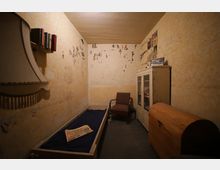 Ein kleiner, schlichter Raum mit gelblichen Wänden, die von verblassten Zeichnungen und Zeitungsausschnitten bedeckt sind. Möbliert mit einem schmalen Bett, einem Sessel, einem Schrank mit Glasfront und einer Holztruhe; an der Wand hängt ein Regal mit Büchern.