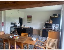 Ein rustikaler Küchen- und Essbereich mit Holzbalken an der Decke, einem Holztisch und Stühlen. Im Hintergrund befinden sich eine schwarze Küchenzeile mit Ofen und Herd, eine weiße Wand mit Fliesen sowie Regale mit Küchenutensilien und Lebensmitteln.