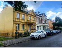 Straßenszene mit historischen Wohnhäusern in einem Gründerzeitstil, gekennzeichnet durch verzierten Fassaden in Gelb und Weiß mit Säulenelementen und Stuckdetails. Im Vordergrund geparkte Autos entlang der gepflasterten Straße, flankiert von einem schmalen Bürgersteig und Eisenzaun.
