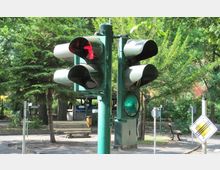 Eine Verkehrsampel mit rot und grün leuchtenden Signalen inmitten einer grünen, baumreichen Umgebung. Im Hintergrund sind Bänke, ein Weg und ein Vorfahrt-gewähren-Schild zu sehen.
