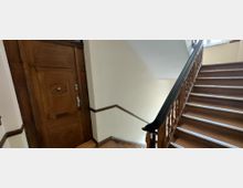 Treppenhaus in einem älteren Wohngebäude mit breiter Holztreppe und geschnitztem Holzgeländer, das zu einem Fenster im oberen Stock führt. Links befindet sich eine massive, verzierte Holztür an hell gestrichenen Wänden mit dunklen Holzleisten.