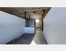 Ein leerer Stall mit weißen, gestrichenen Wänden, rohen Holz- und Betonelementen, Metallgittern und roten Stahlstützen. Ein Fenster im Hintergrund lässt Tageslicht herein.