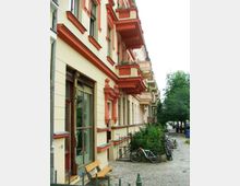 Straßenszene mit Altbaufassaden in hellen Beige- und Rottönen, verziert mit Erkern und Balkonen. Im Vordergrund sind ein gepflasterter Gehweg, Fahrräder an einem Zaun und eine kleine Sitzbank zu sehen, daneben Büsche und Bäume.