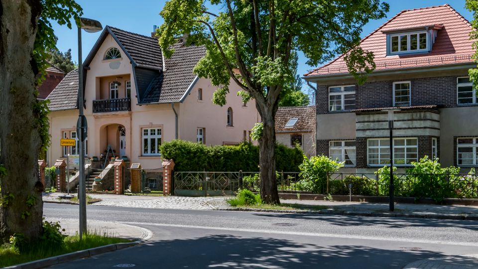 Straßenszene in einem grünen Wohnviertel mit zwei freistehenden Häusern: links ein helles Einfamilienhaus mit steilem Giebeldach und Balkon, rechts ein dunkles Backsteinhaus mit rotem Dach und großen Fenstern. Im Vordergrund verlaufen Straße und Gehweg, eingerahmt von hohen Bäumen, Hecken und niedrigen Zäunen.