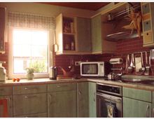 Küche mit mintgrünen Schränken und einer Arbeitsplatte, ausgestattet mit Küchenutensilien, einer Mikrowelle, einem Wasserkocher und einem Herd. Ein Fenster mit kariertem Vorhang lässt Tageslicht herein, während rote Fliesen die Wand hinter der Arbeitsfläche schmücken.