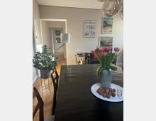 Modernes Wohnzimmer mit Essbereich, einem dunklen Holztisch, darauf eine Vase mit rosa Tulpen und ein Teller mit Walnüssen. Im Hintergrund sind ein heller Holzboden, eine Stehlampe, eine Pflanze und Wanddekoration mit Vintage-Postern zu sehen.