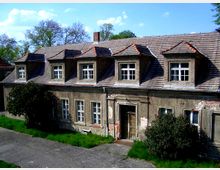 Ein zweistöckiges, altes Gebäude mit bröckelnder Fassade, Satteldach und mehreren Dachgauben; im Vordergrund sind ein gepflasterter Weg, Grasflächen und einige Büsche sichtbar. Im Hintergrund stehen Bäume unter einem blauen Himmel.