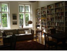 Wohnzimmer mit großen Fenstern, durch die Tageslicht hereinfällt, einer grauen Couch mit Kissen, einem Stehlämpchen und mehreren Bilderrahmen an der Wand. Rechts steht ein großes Bücherregal mit zahlreichen Büchern, daneben ein Tisch mit Stühlen. Der Raum hat einen Holzboden und einen Teppich.