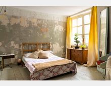 Schlafzimmer mit einem einfachen Holzbett, einer bunten Tagesdecke und Kissen, davor ein abgenutzter Dielenboden. Die Wand ist mit abblätternder Farbe und geometrischen Zeichnungen geziert, ein großes Fenster mit gelben Vorhängen bringt Tageslicht herein, daneben steht eine alte Kommode mit Zimmerpflanzen.