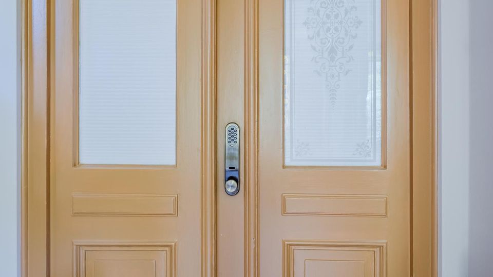 Zweiflügelige Holztür in hellbeigem Anstrich mit profilierten Kassetten und zwei hohen Milchglasfeldern mit ornamentaler Ätzung. In der Mitte befindet sich ein Metallbeschlag mit Zahlencode-Tastatur und runder Griffgarnitur.