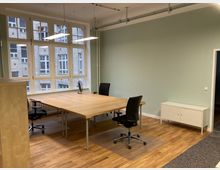 Helles Büro mit großen Fenstern, die auf eine Backsteinfassade blicken. Der Raum verfügt über einen großen Holztisch mit mehreren Bürostühlen, helle Holzfußböden, eine mintgrüne Wand und eine kleine weiße Kommode.