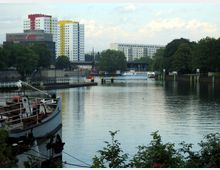 Ein Fluss mit einem festgemachten Schiff im Vordergrund, umgeben von grünen Bäumen und einer Uferpromenade. Im Hintergrund sind moderne Wohngebäude mit farbigen Fassaden sowie eine Brücke und ein vorbeifahrendes Ausflugsschiff sichtbar.