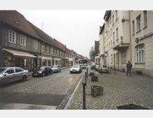 Straßenszene in einer Kleinstadt mit Kopfsteinpflaster, einer Reihe Fachwerkhäuser links und mehrstöckigen Gebäuden rechts. Mehrere parkende und vorbeifahrende Autos sowie ein Linienbus in Fahrtrichtung; Gehwege mit Pollern und leeren Betonblumenkübeln.