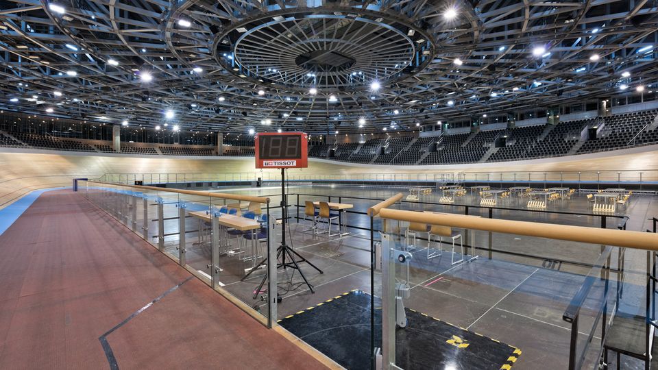 Innenansicht einer modernen Radrennbahn mit ovaler Holzpiste, umlaufenden Tribünen und einem zentralen, ringförmigen Stahl-Dachtragwerk mit vielen Deckenleuchten. Im Vordergrund stehen Glasgeländer, Sitzgelegenheiten und eine digitale Anzeigetafel am Rand der Bahn.