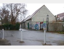 Ein leerer, gepflasterter Platz vor einem alten Gebäude mit schrägem Dach, dessen Wände mit Graffiti bedeckt sind. Rechts steht eine Laterne, im Hintergrund sind kahle Bäume und weitere Gebäude erkennbar.