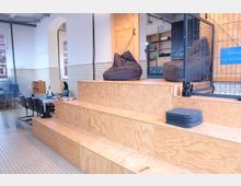 Ein moderner Arbeitsbereich mit Holzelementen, der eine erhöhte Sitzfläche mit Sitzsäcken und gestapelten Kissen aufweist. Im Hintergrund sind Tische mit Stühlen, weiße Fliesenwände, hohe Fenster und Regale erkennbar.