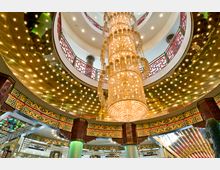 Innenraum mit reich verzierter asiatischer Architektur, einer großen, funkelnden Kristallleuchter im Mittelpunkt und goldenen Deckenpaneelen, umgeben von roten Balustraden mit traditionellen chinesischen Mustern. Die Säulen und Wände sind mit farbenfrohen Ornamenten und typisch asiatischen Designs geschmückt.