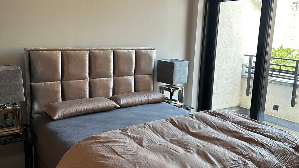 Schlafzimmer mit Doppelbett und gepolstertem Kopfteil, flankiert von zwei Nachttischen mit quadratischen Lampen. Rechts ein großes, schwarz gerahmtes Fenster bzw. eine Balkontür mit Blick auf einen kleinen Balkon mit Metallgeländer.