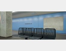 U-Bahn-Station mit hellblauen Wandfliesen, der Schriftzug „Afrikanische Str.“ ist an der Wand zu lesen. Im Vordergrund stehen mehrere schwarze Metallsitze, links ist eine Säule mit kleinen hellen Mosaikfliesen zu sehen.