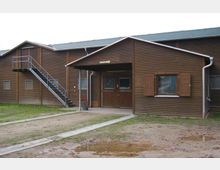 Ein einstöckiges Gebäude aus Holz mit einem kleinen Vordach über dem Eingang, Klappläden an den Fenstern und einer Metall-Außentreppe an der linken Seite. Der Boden davor besteht aus Sand und Gras mit vereinzelten Pfützen.