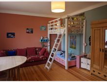 Kinderzimmer mit Etagenbett aus hellem Holz und Leiter, daneben ein rotes Sofa und ein runder Tisch auf Holzdielenboden. Farbige Wände mit Bildern und ein großer Holzschrank prägen den Raum.