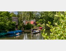 Ein ruhiger Fluss mit einem Kahn voller Menschen, der von einem stehenden Bootsführer gelenkt wird, daneben ein Kajak mit zwei Personen. Im Hintergrund sind grüne Bäume, ein Haus mit Reetdach und ein weiteres mit rotem Ziegeldach zu sehen.