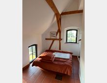 Dachgeschosszimmer mit schrägem, weißem Giebel, sichtbaren Holzbalken und zwei Fenstern mit schwarzen Rahmen. Im Raum steht ein Bett mit rostroter Bettwäsche auf einem Boden mit Terrakottafliesen.