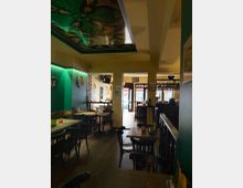 Ein gemütliches Café oder Restaurant mit Holztischen und schwarzen Stühlen, warm beleuchteten Wänden in Grün und Gelb, und buntem Glasdesign an der Decke. Im Hintergrund befindet sich ein Eingangsbereich mit Blick nach draußen sowie eine Bar mit Flaschenregalen.