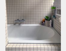 Eine weiße Badewanne mit rechteckigen, weiß-blauen Fliesen an der Wand und um den Wannenrand. Rechts auf dem Rand stehen Pflegeprodukte und eine Topfpflanze.