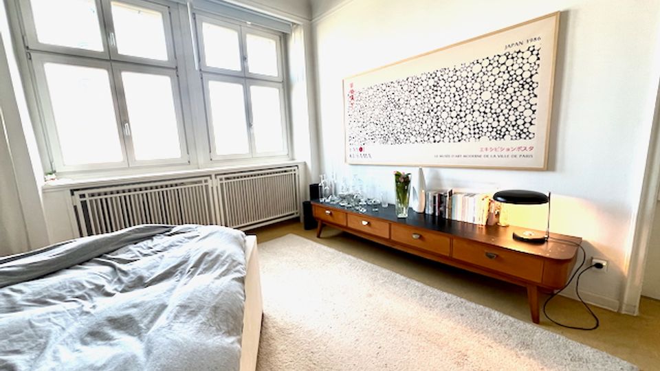 Helles Schlafzimmer mit großen Sprossenfenstern und Heizkörper darunter, einem Bett im Vordergrund und einem langen, niedrigen Sideboard an der Wand. Über dem Sideboard hängt ein großes gerahmtes Wandbild; auf dem Möbel stehen Bücher, Glasflaschen und eine Tischlampe auf hellem Teppichboden.