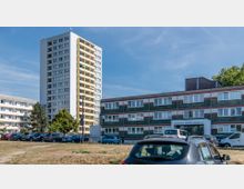 Mehrstöckige Wohngebäude in einer städtischen Umgebung bei Tageslicht; ein Hochhaus mit weißen Balkonen dominiert die Szene, daneben ein niedriges Gebäude mit der Aufschrift "Ärztehaus Prager Straße 35". Im Vordergrund stehen geparkte Autos auf einem trockenen, grasbewachsenen Areal.