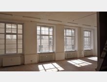 Ein leerer, heller Raum mit vier großen Fenstern, die viel Tageslicht hereinlassen. Die Wände sind weiß gestrichen, der Boden besteht aus Holz, und unter jedem Fenster befindet sich ein Heizkörper.