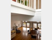 Zweistöckiges Wohnzimmer in einem modernen Haus mit hellem Holzfußboden, großen Fenstern mit Gittern und Blick nach draußen. Im Raum befinden sich ein Sofa, ein Regal, eine Sitzgruppe mit rundem Esstisch und ein Heizkörper an der Wand; oben ist ein weißes Geländer sichtbar.