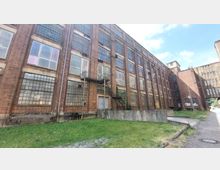 Backsteinfassade eines mehrstöckigen, verlassen wirkenden Industriebauwerks mit großen, rechteckigen Fenstern und Metalltreppen an der Außenseite. Im Vordergrund befinden sich eine Grünfläche und ein gepflasterter Weg, der am Gebäude entlangführt.