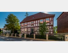 Fachwerkhaus mit weißen und roten Balken, rotem Ziegeldach und Blumen in den Fenstern, umgeben von einem Zaun und einem kleinen Garten. Das Gebäude steht an einer gepflasterten Straße neben anderen Häusern und einem großen Baum.