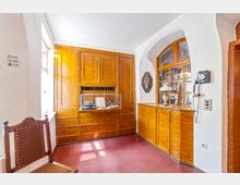 Raum mit historischem Interieur, darunter Wandschränke und Vitrinen aus poliertem Holz sowie ein altes Telefon an der weißen Wand. Eine Vitrine zeigt antike Gegenstände, und der Raum ist durch ein bogenförmiges Fenster mit Holzrahmen erhellt.