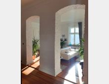 Wohnraum mit sichtbarer weißer Backsteinwand und zwei offenen Bögen, die in ein helles Wohnzimmer führen. Im Wohnzimmer stehen ein cremefarbenes Sofa, ein Tischchen, mehrere grüne Zimmerpflanzen und ein großes Fenster, durch das Tageslicht einfällt.