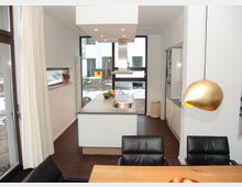 Moderne Küche mit weißer Kochinsel und eingebautem Spülbecken, flankiert von hohen, bodentiefen Fenstern mit Blick nach draußen auf einen schneebedeckten Garten. Im Vordergrund steht ein Esstisch mit schwarzen Lederstühlen und einer goldenen Hängelampe.