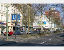 Straßenszene mit Geschäften und einem U-Bahn-Schild für die Linie U9 auf dem Gehweg. Im Hintergrund sind Fassaden von Läden sowie parkende Autos und spärlich belaubte Bäume zu sehen.