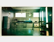 Steriles Untersuchungs- oder Operationszimmer mit Metalltisch in der Mitte, abgedeckt mit einem Tuch, chirurgischer Beleuchtung von der Decke, Arbeitsflächen mit medizinischen Geräten und Schränken im Hintergrund sowie ein Rollhocker und helle Wandlampen.