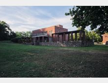 Backsteinbau mit modernem, kubischen Design, umgeben von einer Rasenfläche und Bäumen. Das Gebäude hat einen umlaufenden Balkon, große Fenster und eine offene Pergola auf einer gemauerten Steinsockelstruktur.