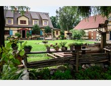 Ein ländlicher Hof mit einem zweistöckigen Fachwerkhaus, gelber Fassade, roten Fensterrahmen und blauen Fensterläden. Im Vordergrund ein Garten mit Rasenfläche, Sitzbereich, Blumenkübeln und einem hölzernen Zaun; rechts ein Nebengebäude mit Holzportalen und einem roten Dach.