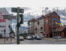Straßenszene in einem städtischen Bereich mit einer Kreuzung; im Vordergrund Verkehr mit Autos und einem Bus, im Hintergrund Gebäude mit verschiedenen Fassadenstilen, darunter ein rotes Backsteinhaus mit großen Fenstern. Straßenschilder und Ampeln sind sichtbar, ebenso wie eine Werbetafel auf der Hauswand links.