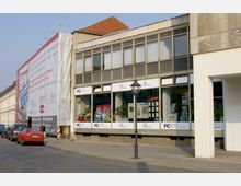 Straßenansicht eines Geschäftsgebäudes mit großflächigen Glasfronten im Erdgeschoss, die ein Reisebüro zeigen. Links des Gebäudes eine Baustelle mit einem Baugerüst und sichtbarer Plane, vor der geparkte Autos stehen.