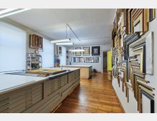 Werkstatt für Bilderrahmen mit hellen Arbeitsflächen, Holzfußboden und an den Wänden lehnenden sowie aufgehängten Rahmenmustern. Im Hintergrund befinden sich Regale mit Materialien, große Fenster mit diffusen Rollos und ein zentraler Werktisch.