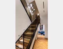 Moderne, helle Inneneinrichtung mit einer Treppe aus Holzstufen und Glasgeländer, die zu einem oberen Stockwerk führt. Im Hintergrund ist ein Wohnbereich mit Parkettboden, blauer Couch und dekorativen Elementen wie einem Tisch und Blumen zu sehen, beleuchtet von einer dekorativen, mehrteiligen Deckenleuchte.