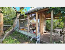 Eine kleine Holz-Gartenhütte mit großen Fenstern, umgeben von Bäumen und Sträuchern. Vor der Hütte befindet sich eine Veranda mit Stühlen und einem Tisch, daneben ein Wasserhahn mit freistehendem Rohr im Grünen.