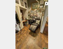 Kleines Tattoo-Studio mit schwarzer Behandlungsliege im Vordergrund, Holzfußboden und Wänden voller Zeichnungen und gerahmter Kunst. Im Hintergrund stehen ein Arbeitstisch mit Hockern sowie Regale und Schubladen mit Tattoo-Utensilien und Lampen.