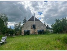 Einfache Hausfassade mit verwittertem Putz und einem Schrägdach, umgeben von einem weitläufigen, grünen Garten mit Bäumen und Sträuchern. Vor dem Haus stehen Gartenmöbel auf einer gepflegten Rasenfläche, und der Himmel ist von dichten Wolken bedeckt.